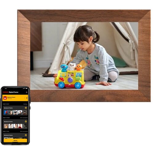 Brązowa ramka cyfrowa wyświetla zdjęcie dziecka bawiącego się kolorową zabawką na podłodze. Obok ramki widoczny jest ekran smartfona z aplikacją "KODAK Digital Frame", pokazującą listę zdjęć i statusy ich wysyłania.
