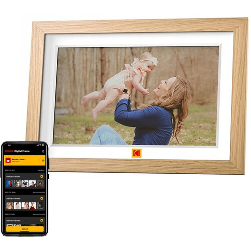 Drewniana ramka cyfrowa z wyświetlaczem pokazującym zdjęcie osoby trzymającej dziecko na tle lasu, z widocznym logo "KODAK" poniżej. Obok ramki znajduje się smartfon z aplikacją "KODAK Digital Frame" pokazującą listę zdjęć do przesłania.