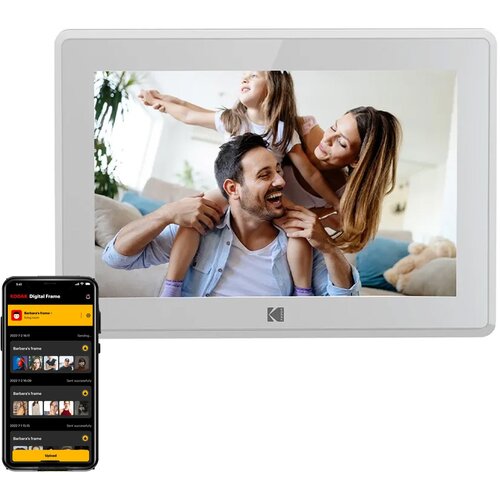 Biała ramka cyfrowa marki Kodak wyświetla zdjęcie trzech osób w domowym otoczeniu. Obok ramki znajduje się smartfon z aplikacją "KODAK Digital Frame", pokazującą listę zdjęć do przesłania.