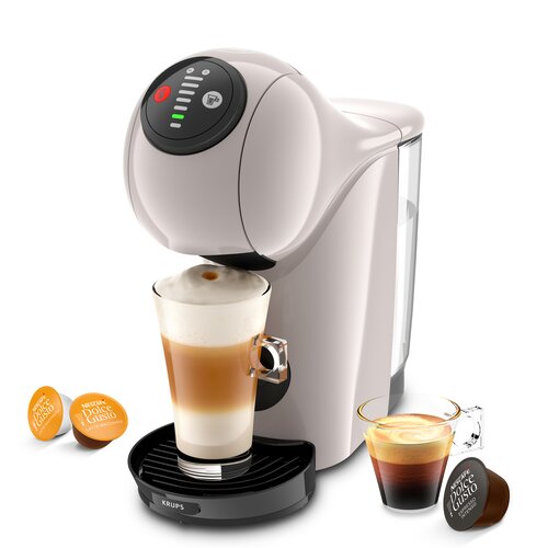 Na zdjęciu znajduje się ekspres do kawy w jasnym kolorze z filiżanką kawy z pianką na podstawce. Obok ekspresu leżą dwie kapsułki z napisem "NESCAFÉ Dolce Gusto" oraz filiżanka z espresso, a na podstawce ekspresu widnieje logo "KRUPS".