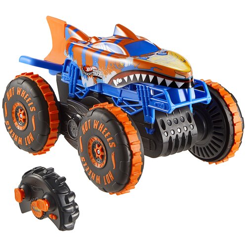 Zdalnie sterowany samochód Hot Wheels Monster Trucks Shark Wspinacz JFR39 z dużymi, pomarańczowymi kołami i niebiesko-pomarańczową karoserią w kształcie rekina, obok znajduje się czarny pilot z pomarańczowymi przyciskami. Na oponach widoczny jest napis "H