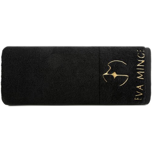 Czarny ręcznik z haftowanym złotym logo i napisem "EVA MINGE" na jednym z końców. Produkt ma teksturę frotte i jest zwinięty w rolkę.