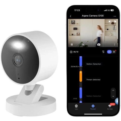 Biała kamera o nowoczesnym designie stoi na podstawce, obok niej znajduje się smartfon z aplikacją wyświetlającą interfejs kamery "Aqara Camera G100". Na ekranie telefonu widoczny jest podgląd z kamery oraz informacje o wykryciu ruchu i osoby, z datą 05/1