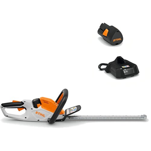 Na zdjęciu widoczne są akumulatorowe nożyce do żywopłotu w kolorze białym i pomarańczowym z czarnymi elementami, z logo "STIHL" na obudowie. Obok znajdują się czarny akumulator oraz ładowarka, również z logo "STIHL".