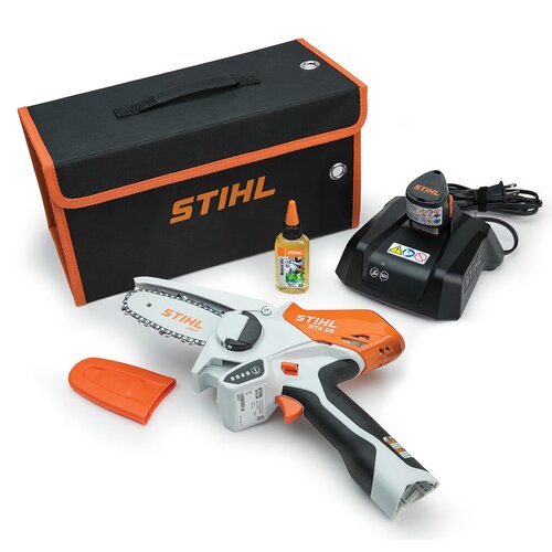 Na zdjęciu znajduje się piła akumulatorowa z widocznym napisem "STIHL GTA 26" na obudowie, obok której leży pomarańczowa osłona na ostrze. W tle widoczna jest czarna torba z pomarańczowym napisem "STIHL", butelka oleju oraz ładowarka z napisem "STIHL" i s