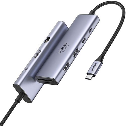 Na zdjęciu widoczny jest hub USB typu C w kolorze srebrnym z kilkoma portami, w tym HDMI i USB, oraz kablem zakończonym wtykiem USB-C. Na urządzeniu znajduje się napis "UGREEN 4K 60HZ".