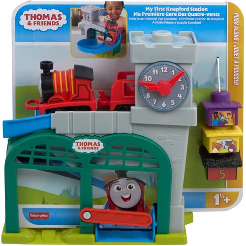 Zestaw zabawkowy przedstawia stację kolejową z czerwonym pociągiem, dużym zegarem i kolorowymi elementami, z widocznymi napisami "THOMAS & FRIENDS" oraz "fisher-price". Opakowanie zawiera teksty w różnych językach, w tym "My First Knapford Station" i ozna