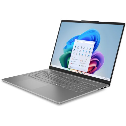 Na zdjęciu widoczny jest otwarty laptop z szarym korpusem, na ekranie którego wyświetlony jest pulpit systemu Windows 11 z ikonami aplikacji. Klawiatura jest pełnowymiarowa, a obudowa ma smukły design.