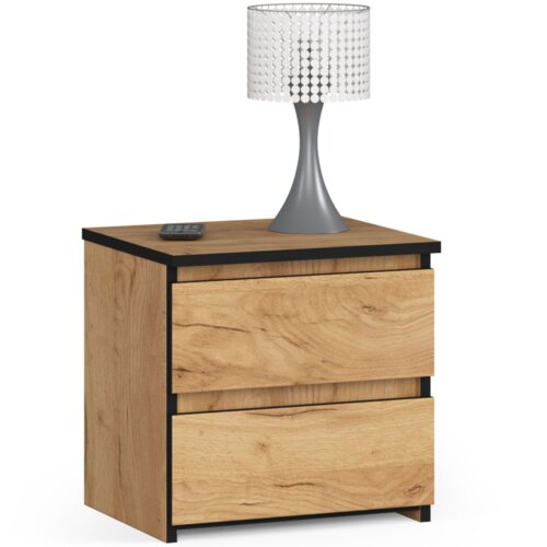 Na zdjęciu znajduje się szafka nocna w kolorze dąb craft z dwoma szufladami, na której stoi lampa z szarym trzonkiem i białym kloszem z ozdobnymi elementami. Obok lampy leży niewielki czarny przedmiot przypominający pilot.