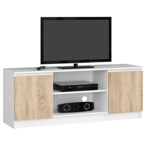 Biała szafka RTV o wymiarach 140x40x55 cm z dwoma drzwiami w kolorze dąb sonoma mat i dwiema półkami, na której stoi telewizor. Na półkach znajdują się czarny odtwarzacz i głośnik.
