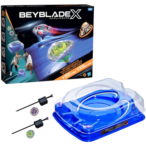 Na zdjęciu znajduje się opakowanie zestawu "Beyblade X" z napisem "Drop Attack" oraz ilustracjami wirujących zabawek i dwóch osób. Obok opakowania widoczna jest niebieska arena do gry oraz dwa wirujące bączki z wyrzutniami.