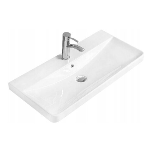 Biała umywalka wpuszczana w blat o prostokątnym kształcie z zaokrąglonymi krawędziami, wyposażona w chromowaną baterię i odpływ. Na powierzchni umywalki widoczne są subtelne refleksy świetlne.