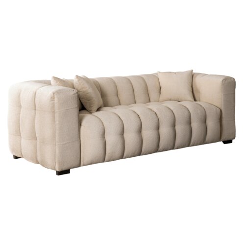 Kremowa, trzyosobowa sofa o pikowanej powierzchni z dwoma poduszkami, ustawiona na ciemnych, prostokątnych nóżkach. Sofa ma elegancki, nowoczesny design z zaokrąglonymi podłokietnikami.