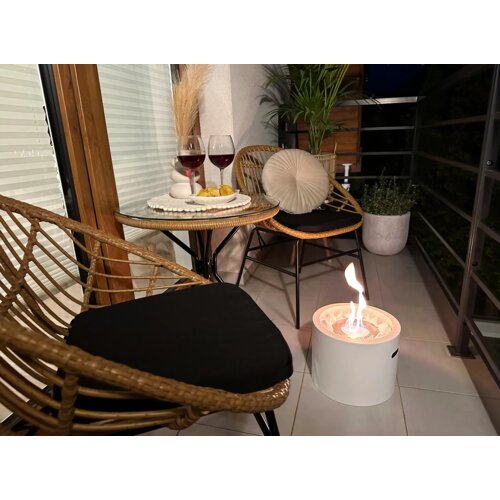 Na zdjęciu znajduje się balkon z dwoma rattanowymi krzesłami z czarnymi poduszkami, okrągłym stolikiem z dwoma kieliszkami czerwonego wina i talerzem z przekąskami oraz białym, wolnostojącym biokominkiem z płomieniem. W tle widoczna jest roślina doniczkowa oraz poduszka dekoracyjna na jednym z krzeseł.